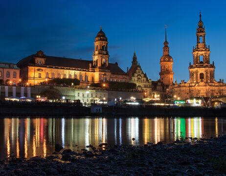 Bunte historische Gebäude und eine Kirche mit grüner Kuppel spiegeln sich in einem ruhigen Fluss unter einem bewölkten Himmel.