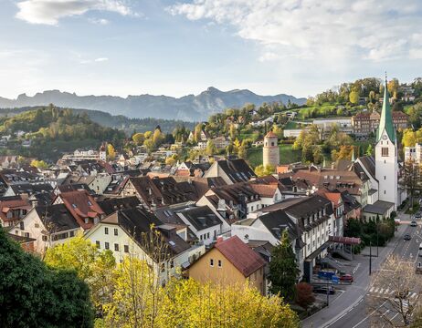 Stadt mit roten Dächern, einer weißen Kirche und Bergen im Hintergrund bei teilweise bewölktem Himmel.