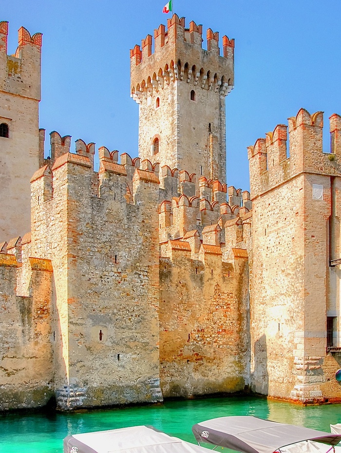 Mittelalterliche Steinburg mit zinnenbewehrten Türmen an türkisfarbenem Wasser unter strahlend blauem Himmel.