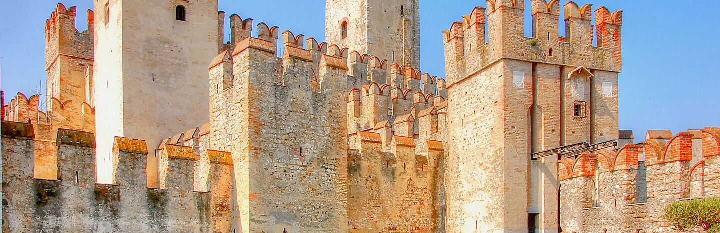 Mittelalterliche Steinburg mit zinnenbewehrten Türmen an türkisfarbenem Wasser unter strahlend blauem Himmel.