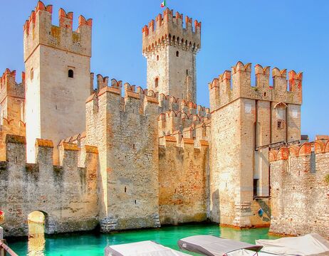 Mittelalterliche Steinburg mit zinnenbewehrten Türmen an türkisfarbenem Wasser unter strahlend blauem Himmel.