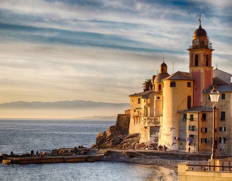 Ein farbenfrohes Gebäude am Meer mit einem Uhrenturm überblickt das ruhige Meer bei Sonnenuntergang, wobei die Berge in der Ferne zu sehen sind.