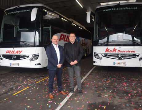 Zwei Männer in Anzügen stehen vor zwei Bussen in einer Garage; buntes Konfetti ist auf dem Boden verstreut.
