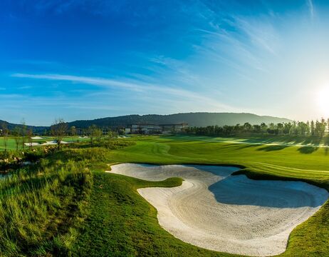 Grüner Golfplatz mit Sandbunker unter strahlend blauem Himmel, Hügeln und einem Gebäude im Hintergrund.
