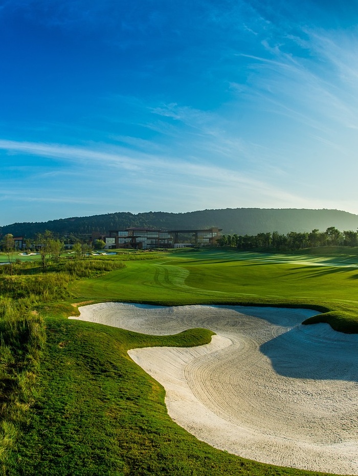 Grüner Golfplatz mit Sandbunker unter strahlend blauem Himmel, Hügeln und einem Gebäude im Hintergrund.