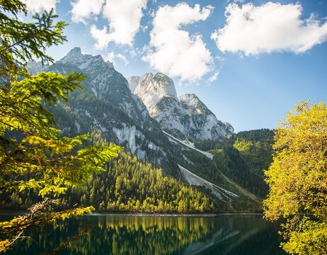 Sonnenbeschienene Berge und Bäume spiegeln sich in einem ruhigen See unter einem blauen Himmel mit vereinzelten Wolken.
