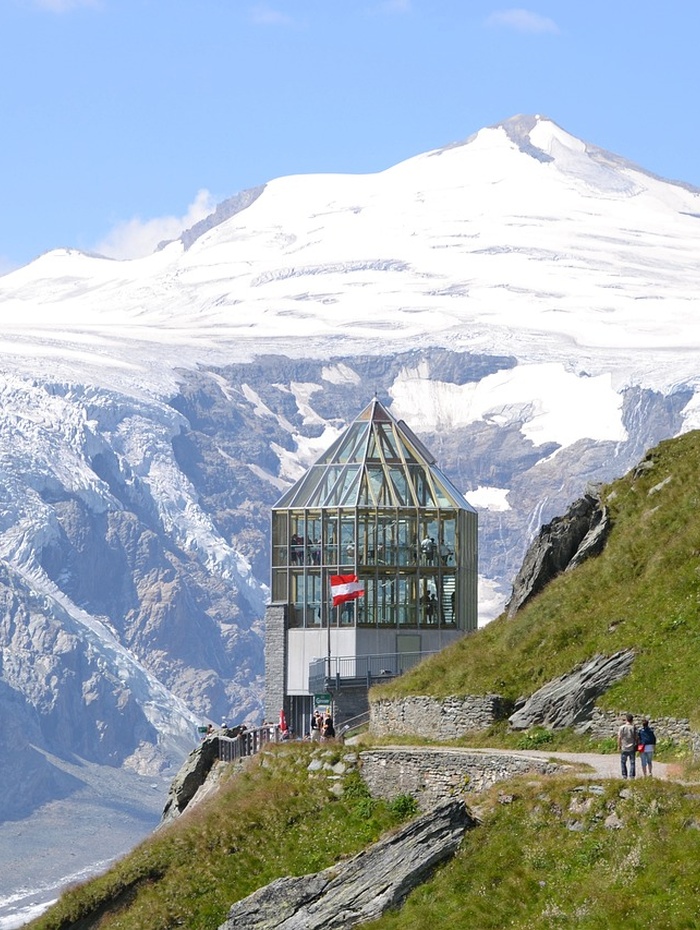 Ein gläsernes Gebäude mit einer Schweizer Fahne liegt auf einem Bergpfad, mit schneebedeckten Gipfeln und Gletschern im Hintergrund.