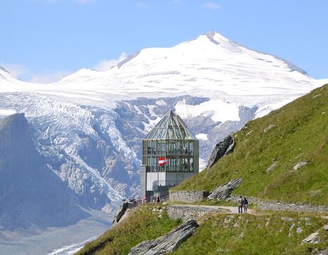 Ein gläsernes Gebäude mit einer Schweizer Fahne liegt auf einem Bergpfad, mit schneebedeckten Gipfeln und Gletschern im Hintergrund.