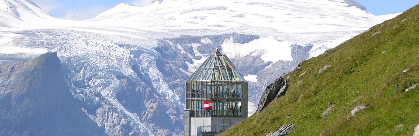 Ein gläsernes Gebäude mit einer Schweizer Fahne liegt auf einem Bergpfad, mit schneebedeckten Gipfeln und Gletschern im Hintergrund.