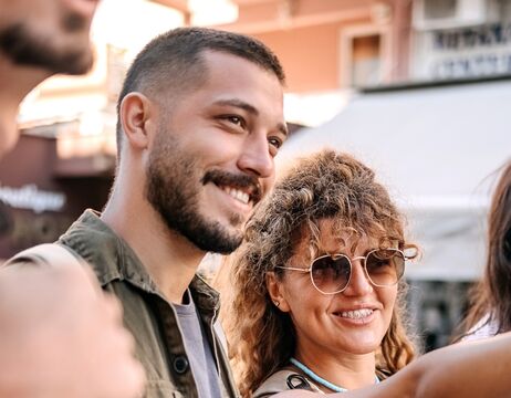 Zwei Menschen lächeln im Freien, einer trägt eine Sonnenbrille, sie wirken glücklich und entspannt in einer lockeren Umgebung.