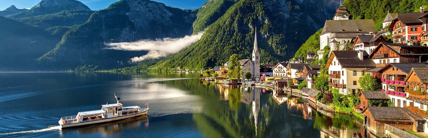 Ein Dorf am See mit einer Kirche, bunten Häusern und Bergen, die sich im ruhigen Wasser unter einem blauen Himmel spiegeln.
