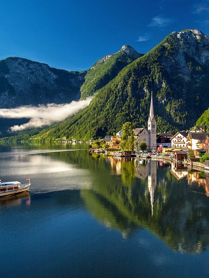 Ein Dorf am See mit einer Kirche, bunten Häusern und Bergen, die sich im ruhigen Wasser unter einem blauen Himmel spiegeln.