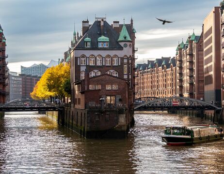Ein historisches Backsteingebäude liegt zwischen Kanälen, mit einem Boot, Brücken und Herbstbäumen im Hintergrund.