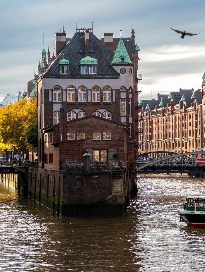 Ein historisches Backsteingebäude liegt zwischen Kanälen, mit einem Boot, Brücken und Herbstbäumen im Hintergrund.