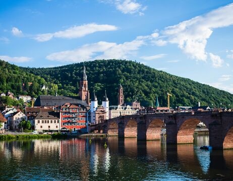 Steinbrücke über einen Fluss mit bunten Gebäuden und einem grünen Hügel im Hintergrund unter blauem Himmel.