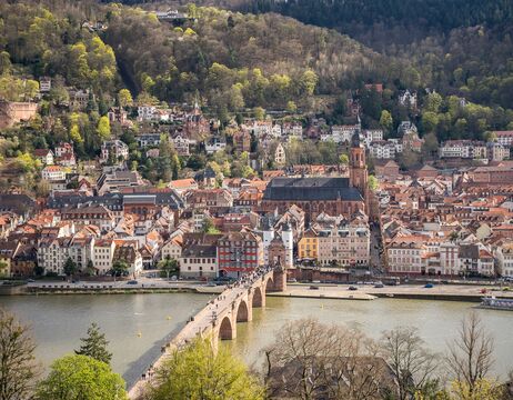 Eine Steinbrücke führt über einen Fluss zu einer historischen Stadt mit roten Dächern und einer Kirche, umgeben von grünen Hügeln.