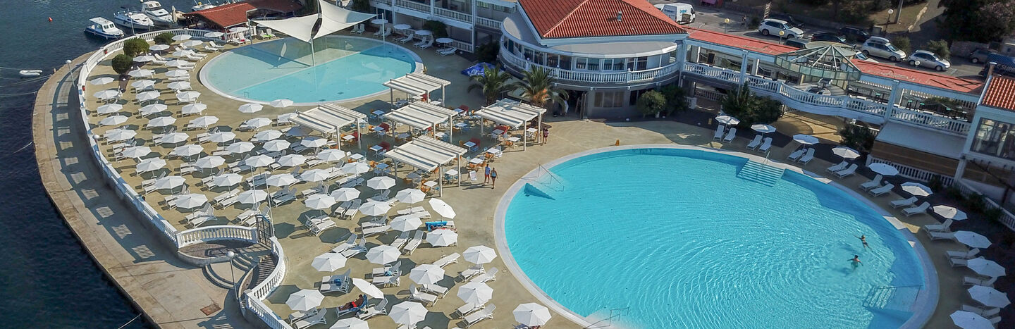 Luftaufnahme eines Badeortes mit großen runden Pools, Liegestühlen und Sonnenschirmen am Wasser.