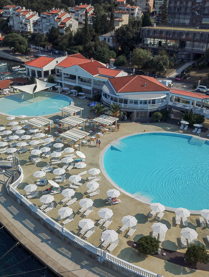 Luftaufnahme eines Badeortes mit großen runden Pools, Liegestühlen und Sonnenschirmen am Wasser.