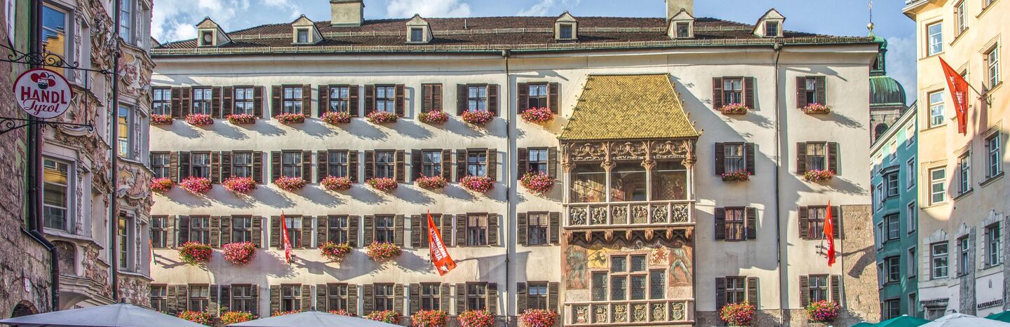 Historisches Gebäude mit vielen Blumenkästen und einem goldenen Balkon an einem belebten Platz unter einem teilweise bewölkten Himmel.