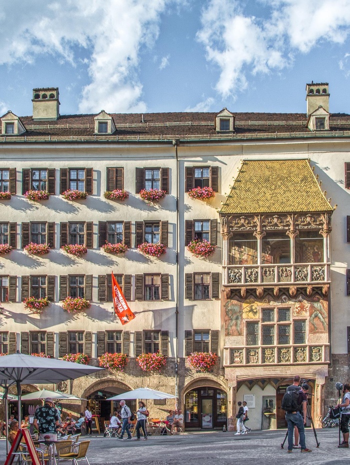 Historisches Gebäude mit vielen Blumenkästen und einem goldenen Balkon an einem belebten Platz unter einem teilweise bewölkten Himmel.