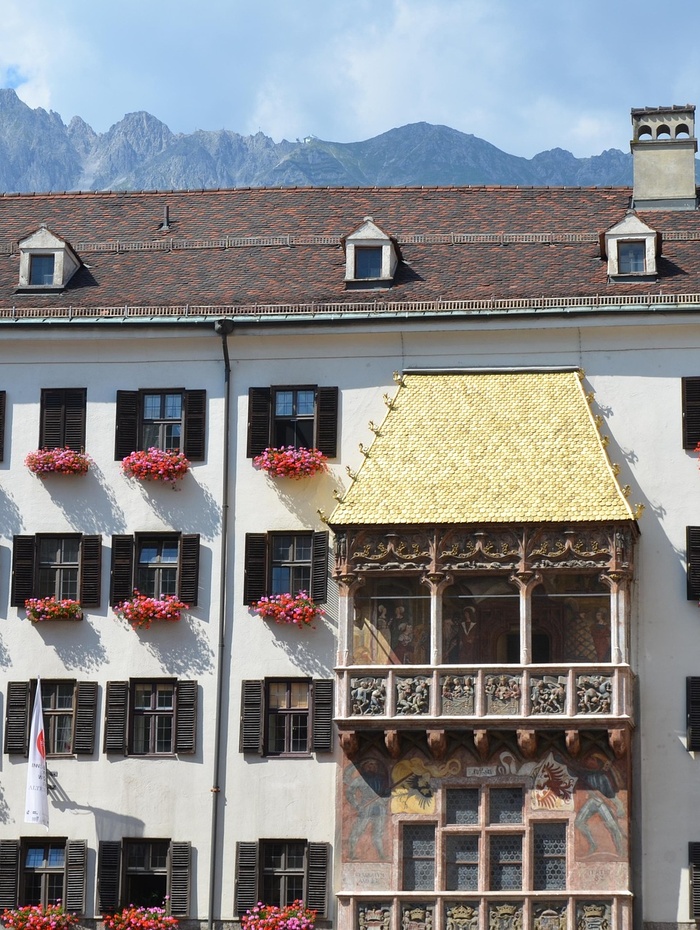 Historisches Gebäude mit einem goldenen Balkon, Blumenkästen und Bergen im Hintergrund.