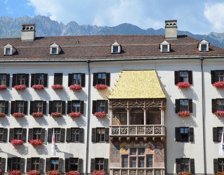 Historisches Gebäude mit einem goldenen Balkon, Blumenkästen und Bergen im Hintergrund.