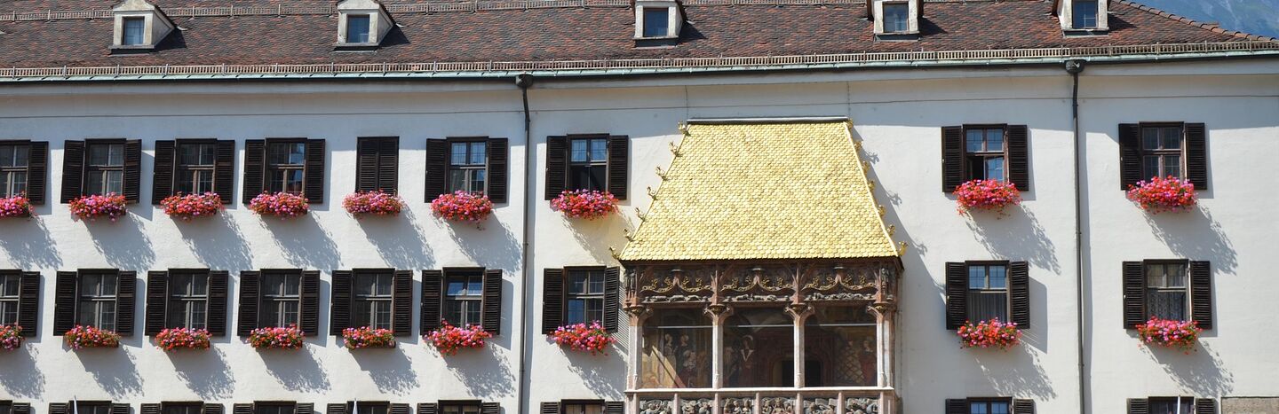 Historisches Gebäude mit einem goldenen Balkon, Blumenkästen und Bergen im Hintergrund.