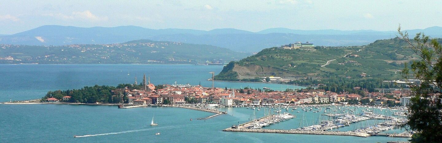 Eine Küstenstadt mit Booten im Yachthafen, umgeben von blauem Wasser und grünen Hügeln unter einem teilweise bewölkten Himmel.