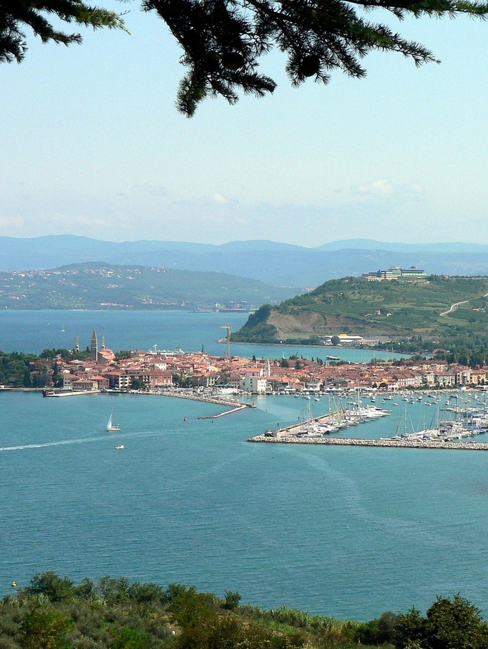 Eine Küstenstadt mit Booten im Yachthafen, umgeben von blauem Wasser und grünen Hügeln unter einem teilweise bewölkten Himmel.