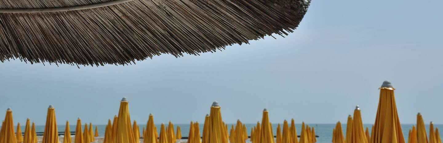 Reihen von geschlossenen gelben Sonnenschirmen und Liegestühlen unter einem großen Strohsonnenschirm am Meer.