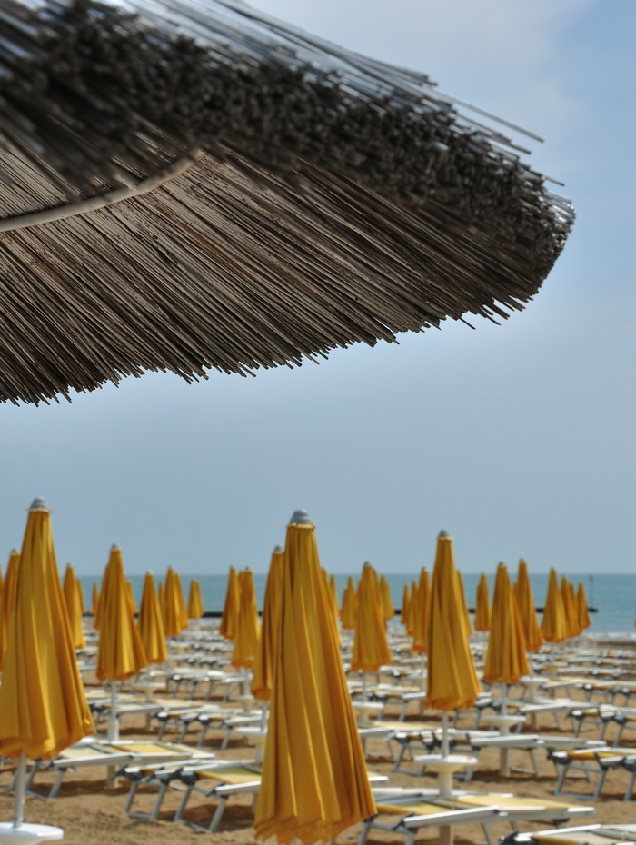 Reihen von geschlossenen gelben Sonnenschirmen und Liegestühlen unter einem großen Strohsonnenschirm am Meer.