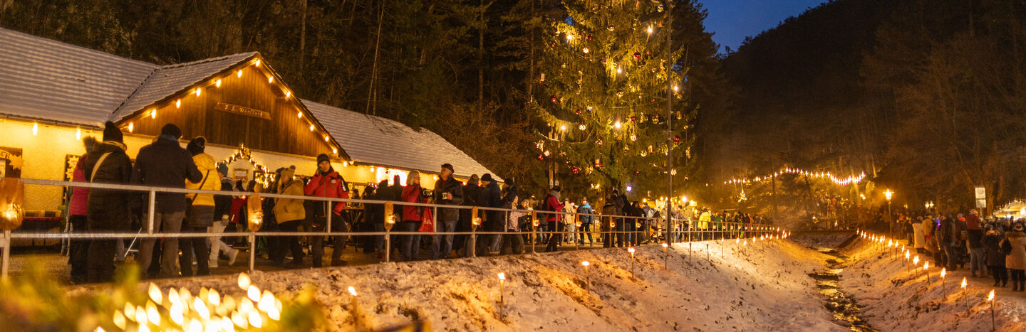 An einem verschneiten Abend versammeln sich die Menschen vor einem großen, geschmückten Weihnachtsbaum und festlichen Lichtern.