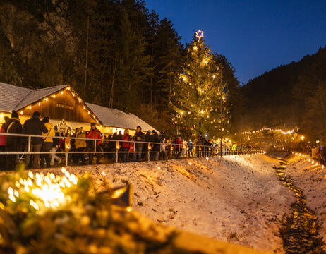 An einem verschneiten Abend versammeln sich die Menschen vor einem großen, geschmückten Weihnachtsbaum und festlichen Lichtern.