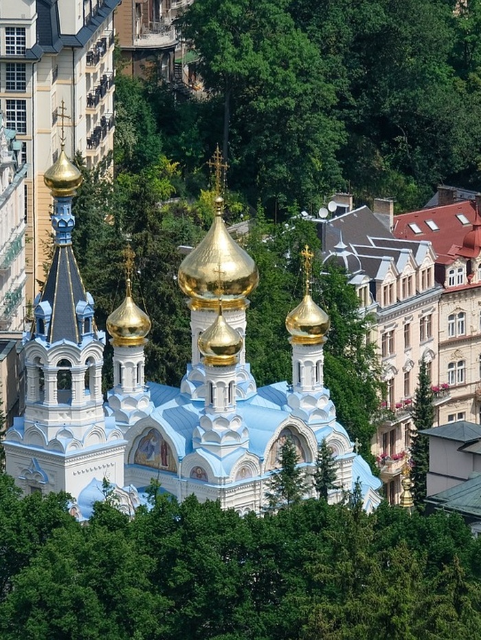 Weiß-blaue Kirche mit goldenen Kuppeln, umgeben von Bäumen und städtischen Gebäuden im Hintergrund.