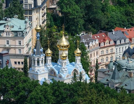 Weiß-blaue Kirche mit goldenen Kuppeln, umgeben von Bäumen und städtischen Gebäuden im Hintergrund.
