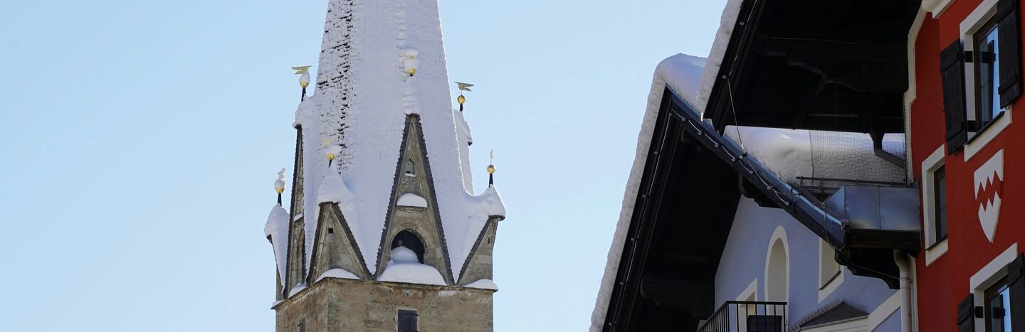 Der schneebedeckte Kirchturm erhebt sich über den farbenfrohen Alpengebäuden unter einem klaren blauen Himmel.