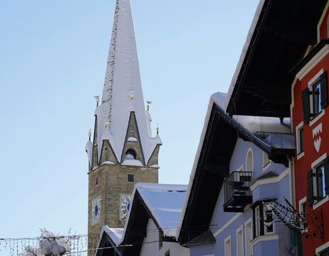 Der schneebedeckte Kirchturm erhebt sich über den farbenfrohen Alpengebäuden unter einem klaren blauen Himmel.