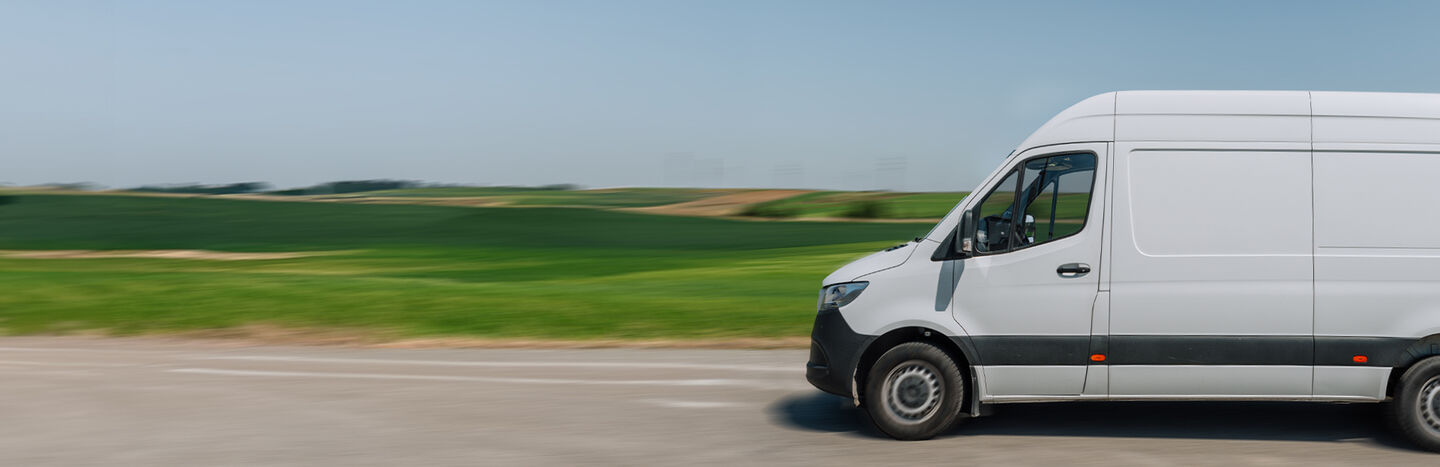 Weißer Transporter auf einer Landstraße mit grünen Feldern und klarem blauen Himmel im Hintergrund.