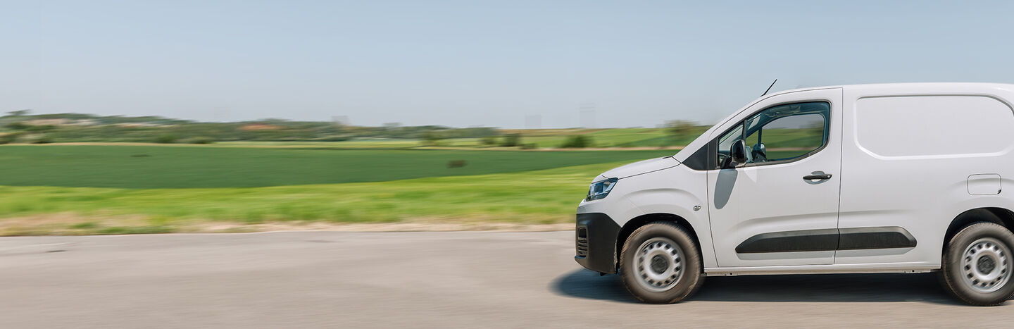Ein weißer Lieferwagen fährt auf einer Straße mit grünen Feldern und blauem Himmel im Hintergrund.