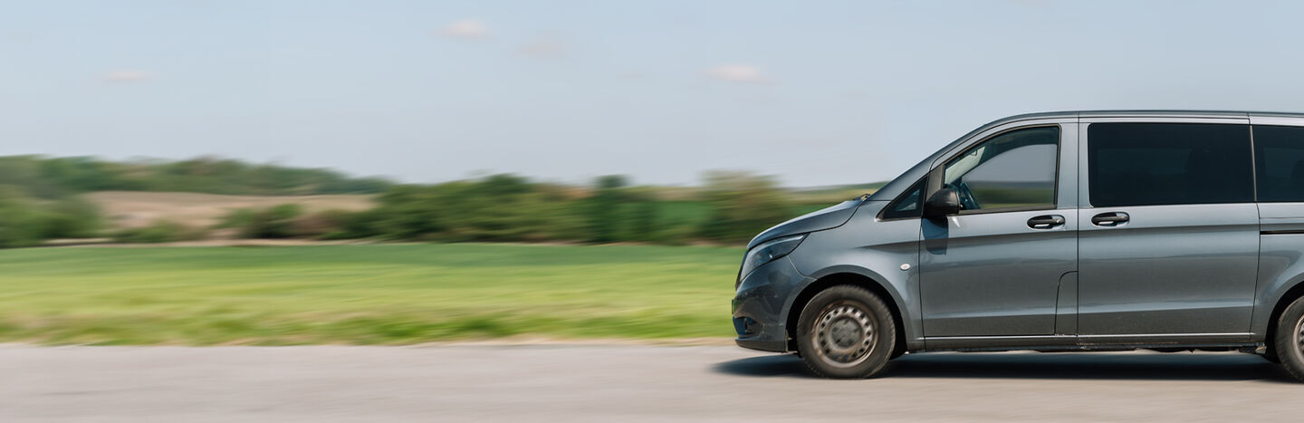 Ein grauer Lieferwagen fährt schnell auf einer Landstraße mit grünen Feldern und Bäumen im Hintergrund.