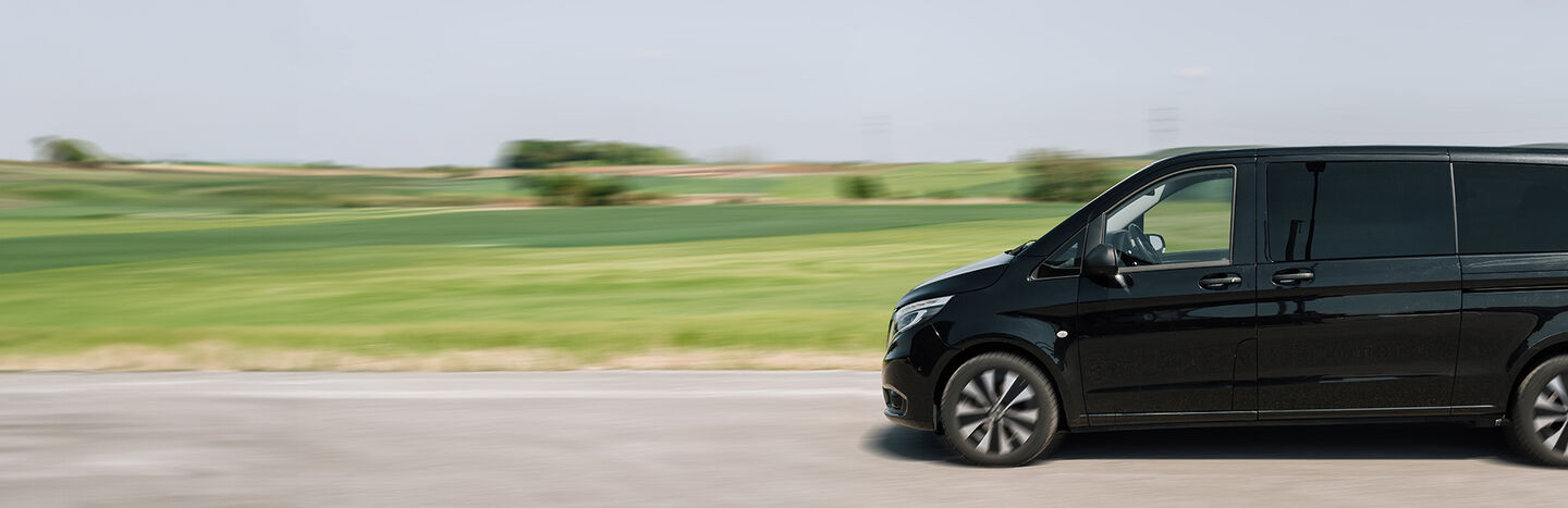 Ein schwarzer Lieferwagen fährt auf einer Straße mit grünen Feldern und einem klaren Himmel im Hintergrund.
