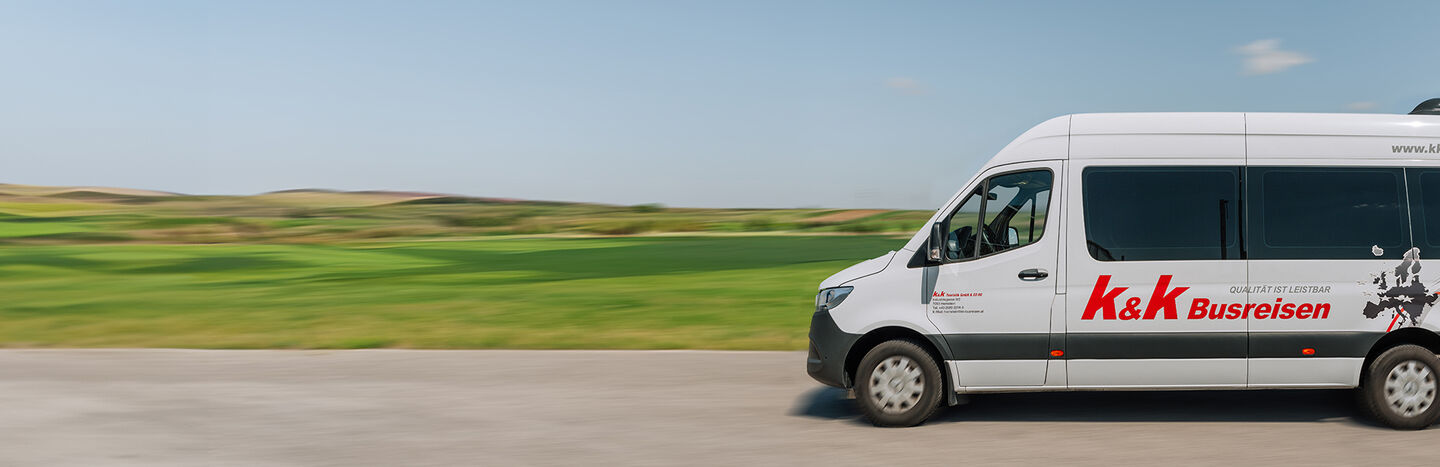 Ein weißer Transporter von K&K Buseisen fährt auf einer Straße mit grünen Feldern und Hügeln im Hintergrund.