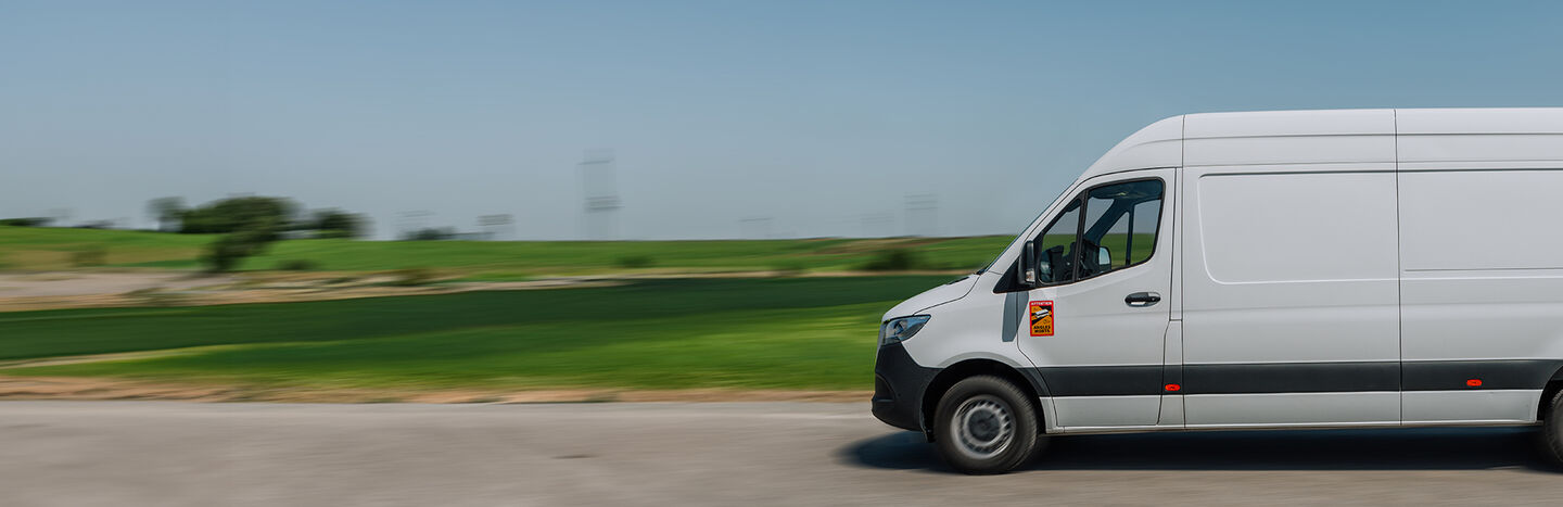 Weißer Lieferwagen fährt schnell auf einer Landstraße mit grünen Feldern und blauem Himmel im Hintergrund.