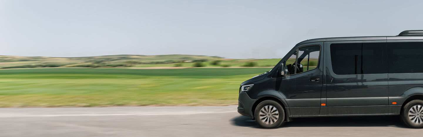 Ein schwarzer Lieferwagen fährt auf einer Straße mit grünen Feldern und Hügeln im Hintergrund unter einem klaren Himmel.