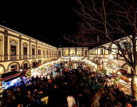 Ein festlicher Nachtmarkt erstrahlt mit Lichtern und Menschenmassen in einem historischen, von Säulen gesäumten Innenhof.
