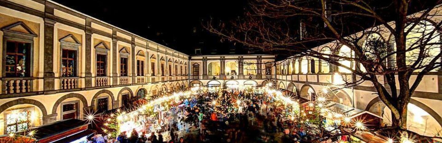 Ein festlicher Nachtmarkt erstrahlt mit Lichtern und Menschenmassen in einem historischen, von Säulen gesäumten Innenhof.