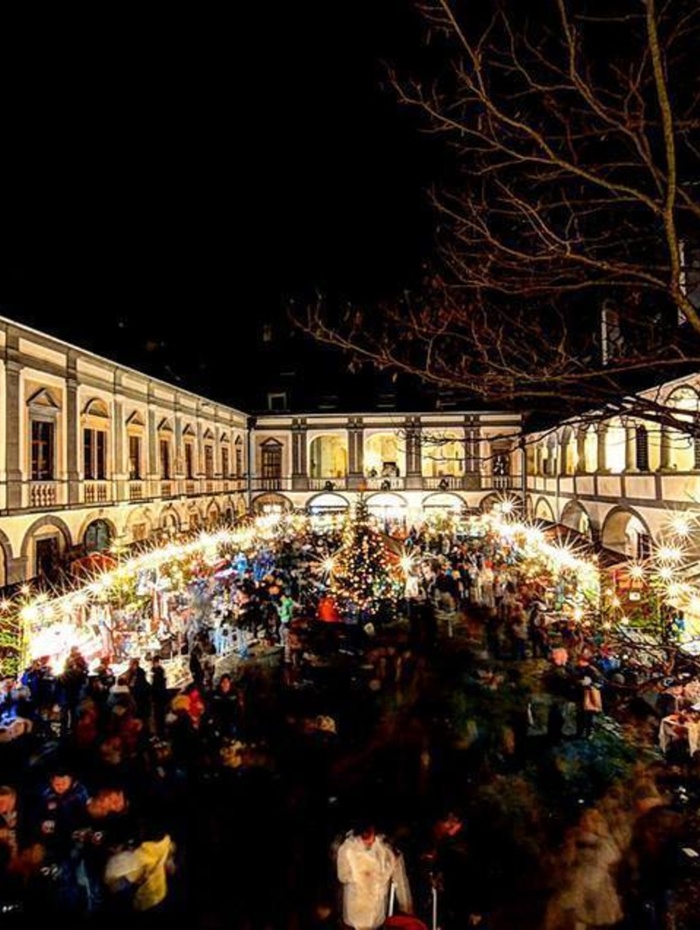 Ein festlicher Nachtmarkt erstrahlt mit Lichtern und Menschenmassen in einem historischen, von Säulen gesäumten Innenhof.