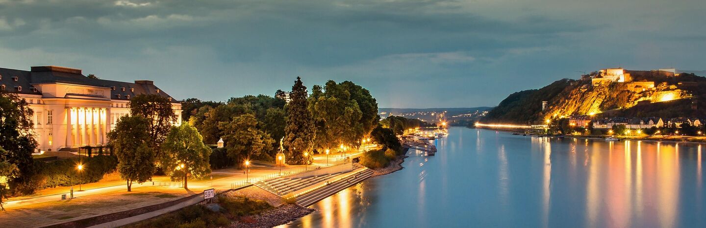 Ein ruhiger Fluss in der Abenddämmerung mit Straßenlaternen, die sich auf dem Wasser spiegeln, und Bäumen am Ufer.
