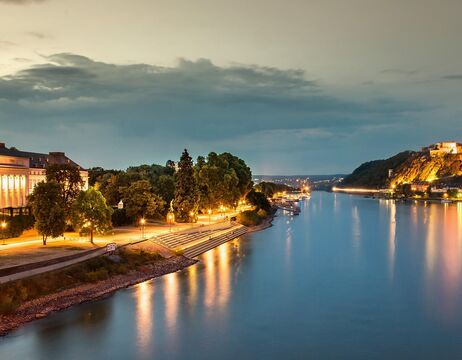 Ein ruhiger Fluss in der Abenddämmerung mit Straßenlaternen, die sich auf dem Wasser spiegeln, und Bäumen am Ufer.