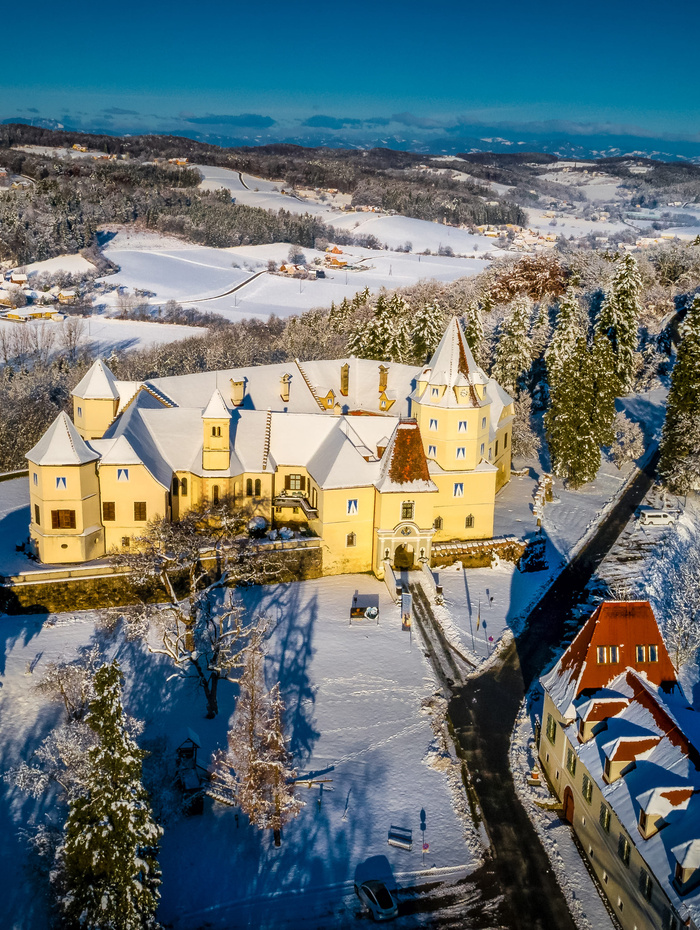 Luftaufnahme eines gelben Schlosses und einer verschneiten Landschaft mit Bäumen, Hügeln und nahe gelegenen Gebäuden unter einem klaren blauen Himmel.